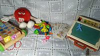 Toy desk with magnetic letters, pink chalk, instruction cards, M&M plush toy, and wooden pull-along block toy on patterned fabric background.