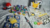 Wide shot showing the full Pokémon lot including cards, figurines, lunchbox, game components, and wristwatch arranged on a lace tablecloth