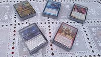 Five separate stacks of Magic The Gathering cards sealed in plastic sleeve, showing a variety of card faces in different colors and styles.