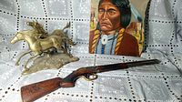 Photo showing miniature double-barrel shotgun replica with carved wooden stock, brass mustangs sculpture, and Native American portrait on quartz slab in background.