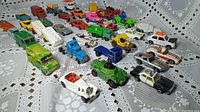 Front angled view of the collection of 32 vintage toy cars on a lace tablecloth, showing variety in color and model type