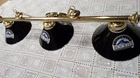 Photo showing full length of brass bar holding three black metal lampshades with Colorado Rockies logos.