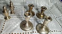 Four sterling silver weighted candlestick holders and two pairs of sterling silver weighted salt and pepper shakers displayed on a lace placemat, showing patina and wear.