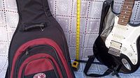 Electric guitar with white pickguard and black body shown next to a tape measure for size reference, along with soft carry backpack bag on the ground.