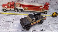 Photo showing both the vintage metal semi fire truck and the black Tonka truck with roll bar on a patterned surface with tape measure for scale indicating the fire truck is about 3 feet long.
