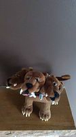 Full side view of the brown plush three-headed dog showing all three heads with white spikes on collars and four detailed legs with claws.