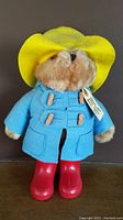 Front view of plush Paddington Bear wearing yellow rubber rain hat, blue coat with wooden toggles, red rubber boots, and attached tag.