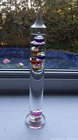 Front view of 12 inch tall glass Galileo thermometer with colorful liquid filled spheres floating inside clear glass cylinder.
