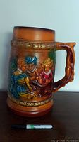 Front view of large ceramic tankard with painted raised relief of three people drinking, displayed next to a Sharpie marker for size reference.