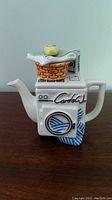 Front view of teapot showing washing machine face, signature, and laundry design.