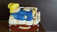 Side view of Paddington Bear cookie jar showing bear in blue coat and yellow hat lying on biscuits in open brown suitcase