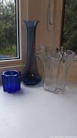 Photo showing the three glass items on a windowsill: clear ruffle handkerchief vase, tall blue vase, blue star votive holder.