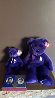 Two purple Ty Beanie Bears, one larger Beanie Buddy and one smaller Beanie Baby, with a rectangular tin of playing cards showing portraits of Prince Charles and Princess Diana.