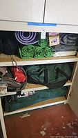 Multiple rolled sleeping pads, tarps, Coleman pump and black stuff sacks on a shelf