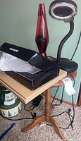 Photo of the wooden round corner table with three lamps on top: a red lava lamp, a desk lamp with circular glass shade, and a box with packaging material, showing the table legs and electrical cords on the floor.