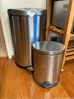 Two stainless steel garbage cans side by side. Larger can is about 27 inches. Blue tape on large can's body.