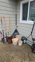 Overall display of gardening tools including shovels, a rake, hand tool, plant pots, watering can, metal planter box, and a push mower.