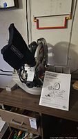 Front view of Craftsman miter saw showing dust collection bag with Craftsman branding and operator's manual beside it.
