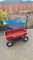 Wide view of blue Jackson wheelbarrow and red Radio Flyer Big Red Classic ATV wagon on outdoor cement surface with wooden fence background