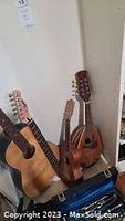 Three string instruments upright against wall: two six-string acoustic guitars in natural wood finish and one smaller four-string ukulele
