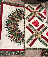Photo showing two quilts: one with a wreath pattern featuring green leaves on white fabric trimmed with red and green holly border, and another with large poinsettia flowers and patterned red and green border.