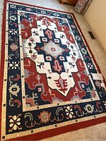 Full view of the rug showing the geometric and floral traditional Indian design with a red, navy and off-white color palette on carpeted floor.