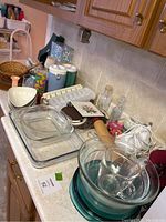Multiple kitchenware items arranged on countertop, including glass bowls, measuring cup, rolling pin, hand mixer, plastic containers, and recipe card boxes.