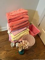 Stack of folded pink, coral, and yellow towels with one pink rectangular waste bin behind. White cup and toothbrush holder, blue ceramic vase with faux pink flowers in front on wood floor.