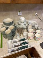 Overview of assorted dishes including green gradient bowls and mugs, white plates and bowls with green trim, floral mugs, cream and sugar set, green-handled cutlery, and decorated juice glasses.