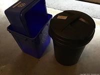 Photo showing black plastic garbage can with attached lid and two blue plastic recycling bins with open hinged lids stacked together on garage floor.