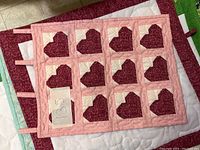 Quilt with 15 dark red hearts on pink patchwork squares bordered in pink fabric.