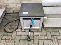 Closed view of beige Suncast hose storage box with grey lid, attached black hose and blue spray nozzle visible outside.