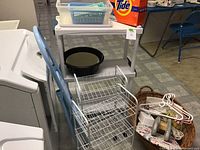 View of white plastic shelving unit, metal wire rolling shelves, blue step ladder, a black basin, and a basket with hangers and laundry accessories.