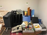 Photo showing a large black electric calculator, black paper shredder, multiple magazine holders, tape dispenser, and various small office supplies arranged on a table.