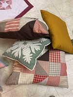 Four quilted pillows on floor, three with patchwork designs in red, beige, brown, and green, one solid mustard yellow.