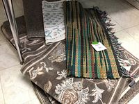 Lot of seven small decorative rugs stacked, showing a variety of colors and patterns including florals and stripes.