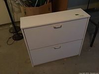 White two-tier shoe organizer cabinet on a concrete floor, showing two drop-down compartments and metal handles.