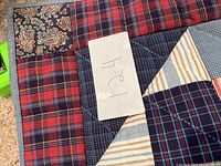 Close-up of handmade patchwork square quilt with plaid, striped, and paisley fabrics showing machine stitched quilting patterns and size tape measure.