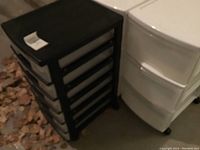 Group view of one black plastic storage drawer unit and one white plastic storage drawer unit side-by-side on garage floor, with dry leaves nearby.