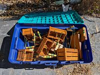 Photo showing multiple small wooden crates or containers with attached wire handles and painted wooden grips inside a blue plastic bin with green lid.