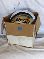 Close-up of Nash hubcap inside partially damaged cardboard box, showing logo and condition.