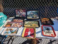 Photo showing the full set of 10 Coca-Cola collectible tin serving trays arranged on a table outdoors. Each tray has different vintage or nostalgic Coca-Cola related illustrations or advertising images.