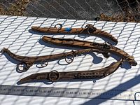 Four primitive antique horse hames laid on wire mesh table outdoors with shadows. Visible iron rings and hardware on aged wooden pieces.