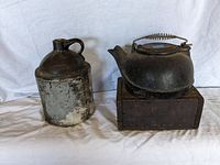 Front view of a weathered ceramic jug and a cast iron teapot glued to a wooden box base on a white cloth background.