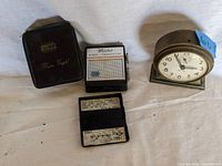 Four vintage items: Mikro First Transistor Radio with case, brass desk clock, Big Ben alarm clock, and Meerschaum box, laid out on a white cloth background.