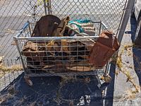 Metal wire mesh cart filled with folded leather chaps and bags, some items hanging off the side. Cart is outdoors, chain link fence background, asphalt ground.