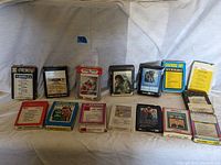 Wide view of sixteen vintage 8-track tapes arranged in two rows on a white sheet background. Various colorful labels and cartridge cases shown.