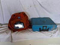 Photo of an orange fabric protective helmet with a flip-up viewing lens sitting next to a blue plastic carrying case on a white surface.
