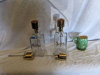 Two rectangular glass decanters labeled 'Vodka' and 'Scotch' with metal pour spout caps, positioned side by side with a small green and cream ceramic cup to the right.