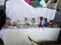 Full view of the lot displaying 10 vintage oil lamps arranged in a row, seven glass lamps and three electrified metal lantern lamps, on a white draped surface.